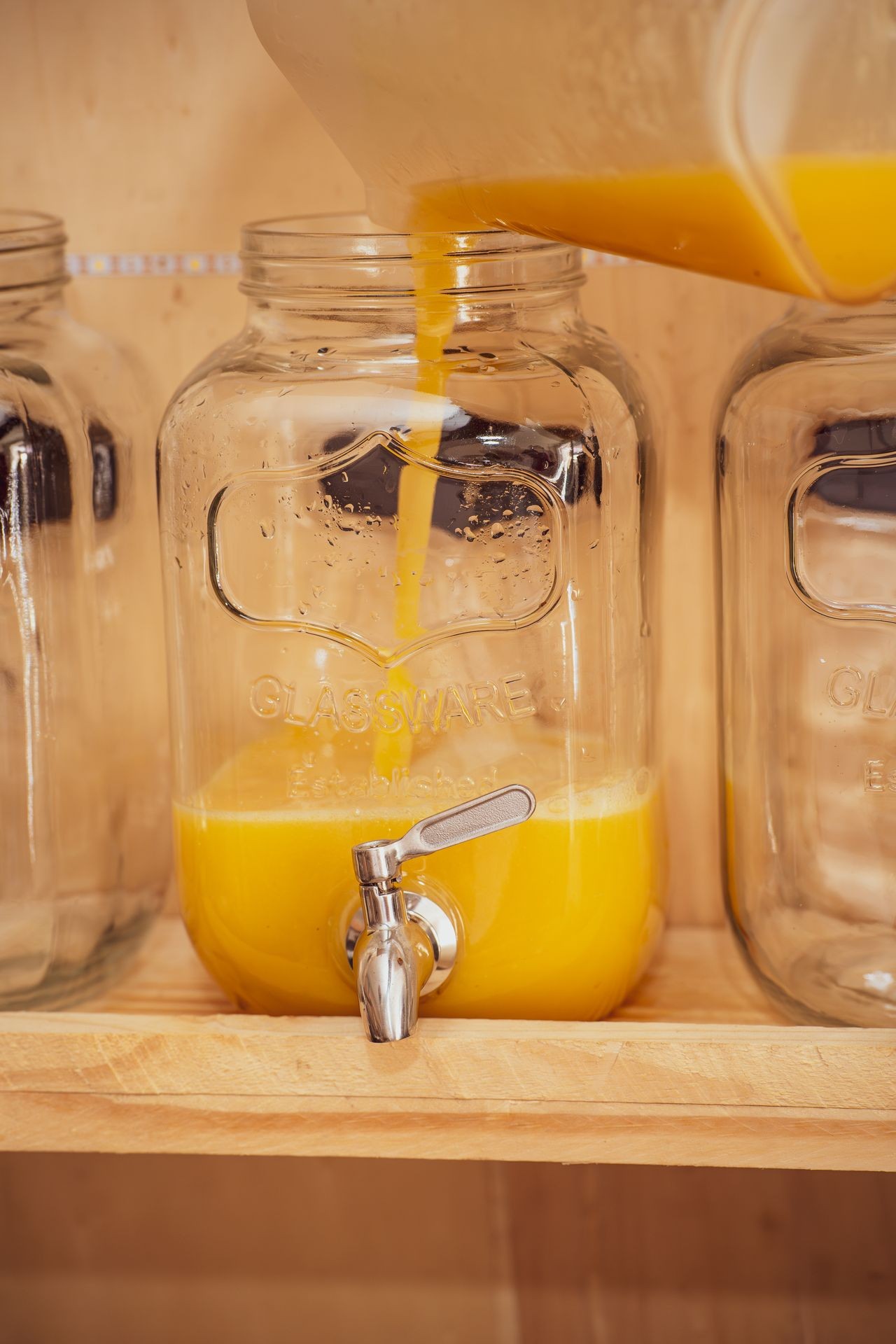Orange juice being poured into a glass dispenser with a spigot on a wooden shelf.