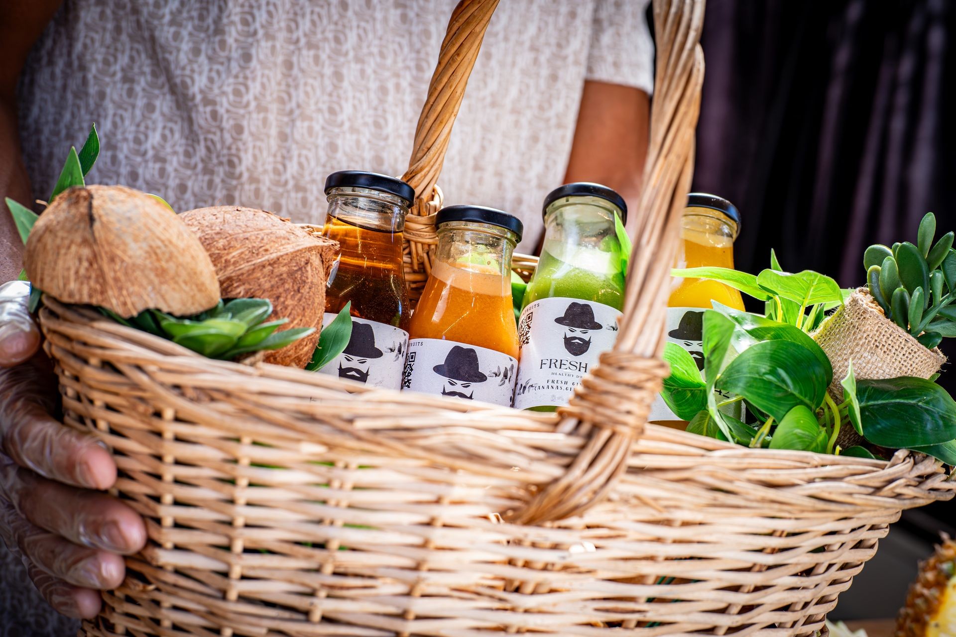 Person holding a wicker basket containing fresh juice bottles, coconuts, and green plants.