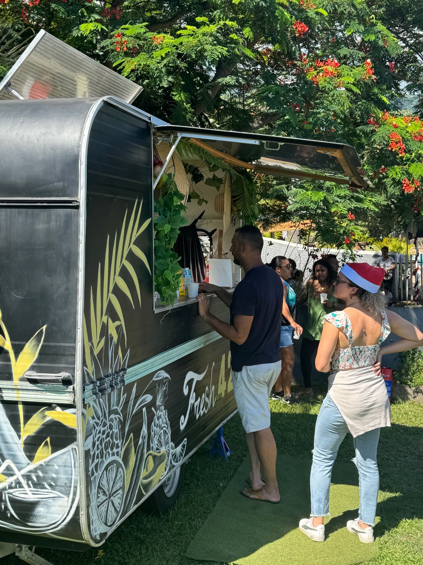 People ordering food from a tropical-themed food truck under lush trees with red flowers.